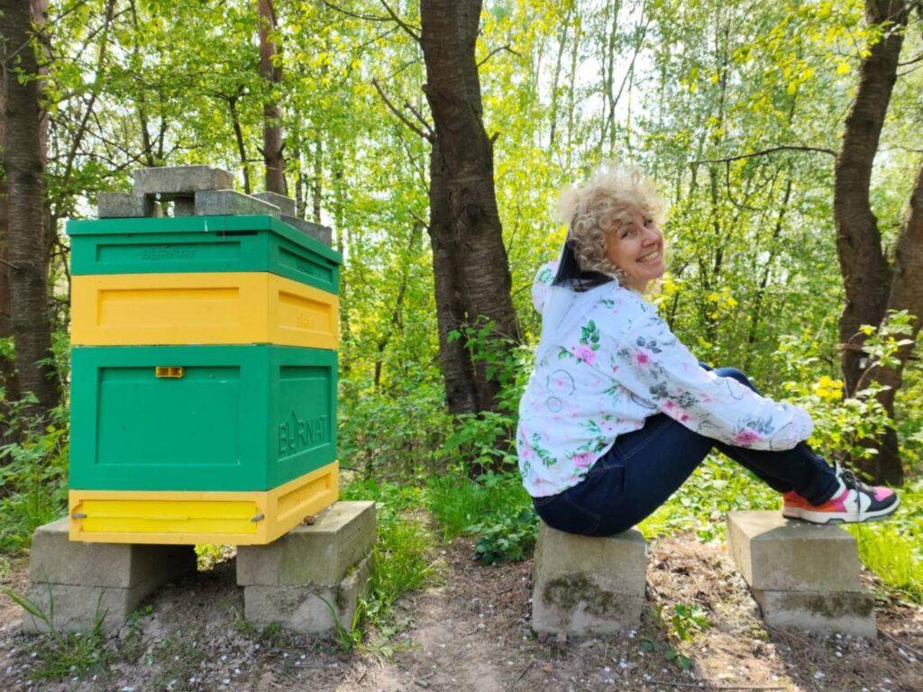 Pszczelarka z Wśród Pszczół i Paproci w kolorowej bluzie pszczelarskiej, siedząca obok ula.